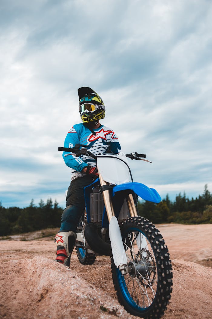 Motorcyclist in blue gear on a dirt bike in an open sandy area with cloudy sky.