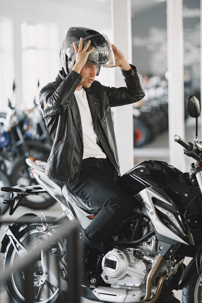 A young man in a leather jacket putting on a helmet while sitting on a motorcycle indoors.